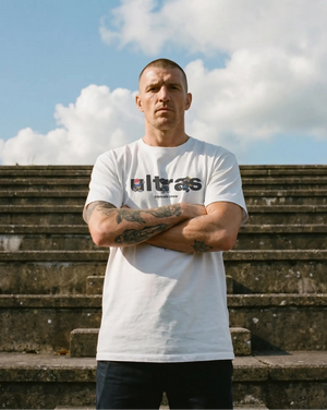 Man wearing a white t-shirt with 'Ultras' logo standing on stone steps under a blue sky with clouds.
