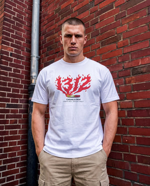 A man wearing a white t-shirt with a red 1312 and black graphic print, standing against a red brick wall.