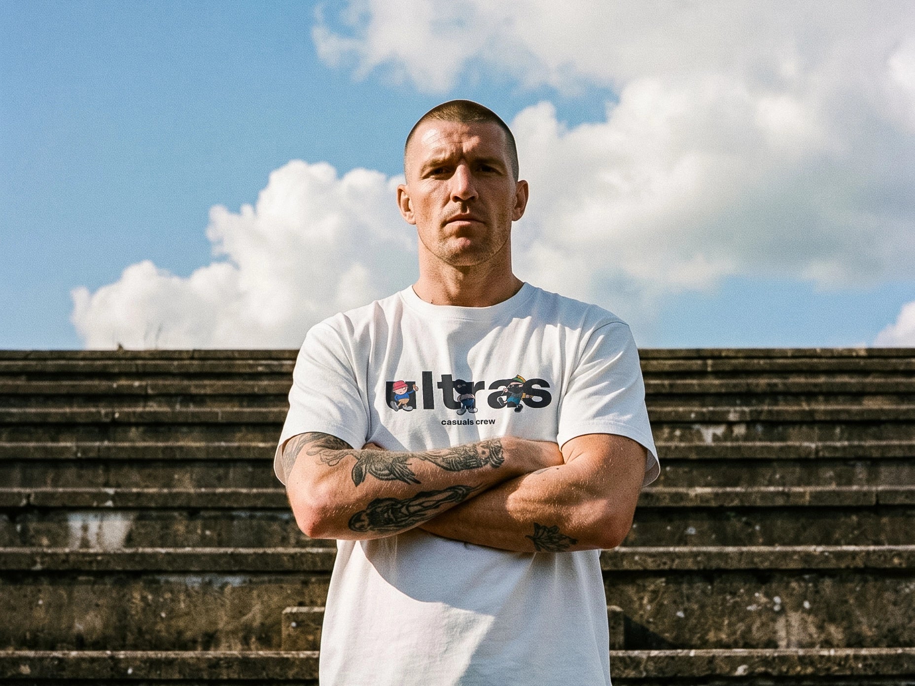 Man wearing a white t-shirt with 'Ultras' logo standing on steps with a cloudy sky background