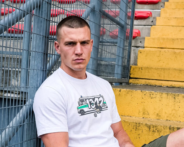 A man sitting on yellow stadium steps in front of a metal fence, wearing a white crewneck t-shirt with a graphic of a truck carrying a '1312' sign, and olive green cargo shorts.