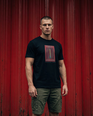 A man stands in front of a red corrugated metal wall wearing a black t-shirt with a red and gold graphic print that reads NO PYRO NO PARTY paired with olive green cargo shorts.