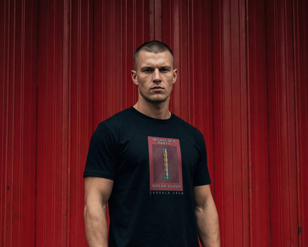 A man stands in front of a red corrugated metal wall wearing a black t-shirt with a red and gold graphic print that reads NO PYRO NO PARTY paired with olive green cargo shorts.